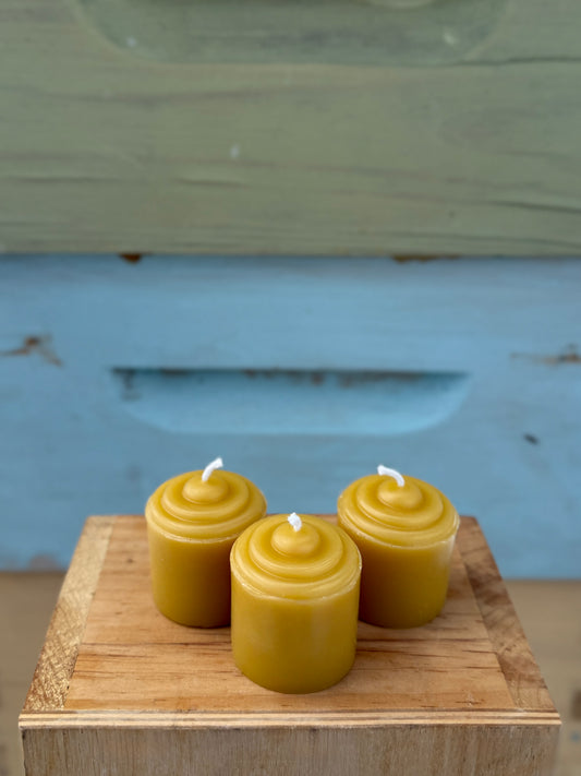 Three yellow beeswax votive candles on a wooden block with a beehive in the background.